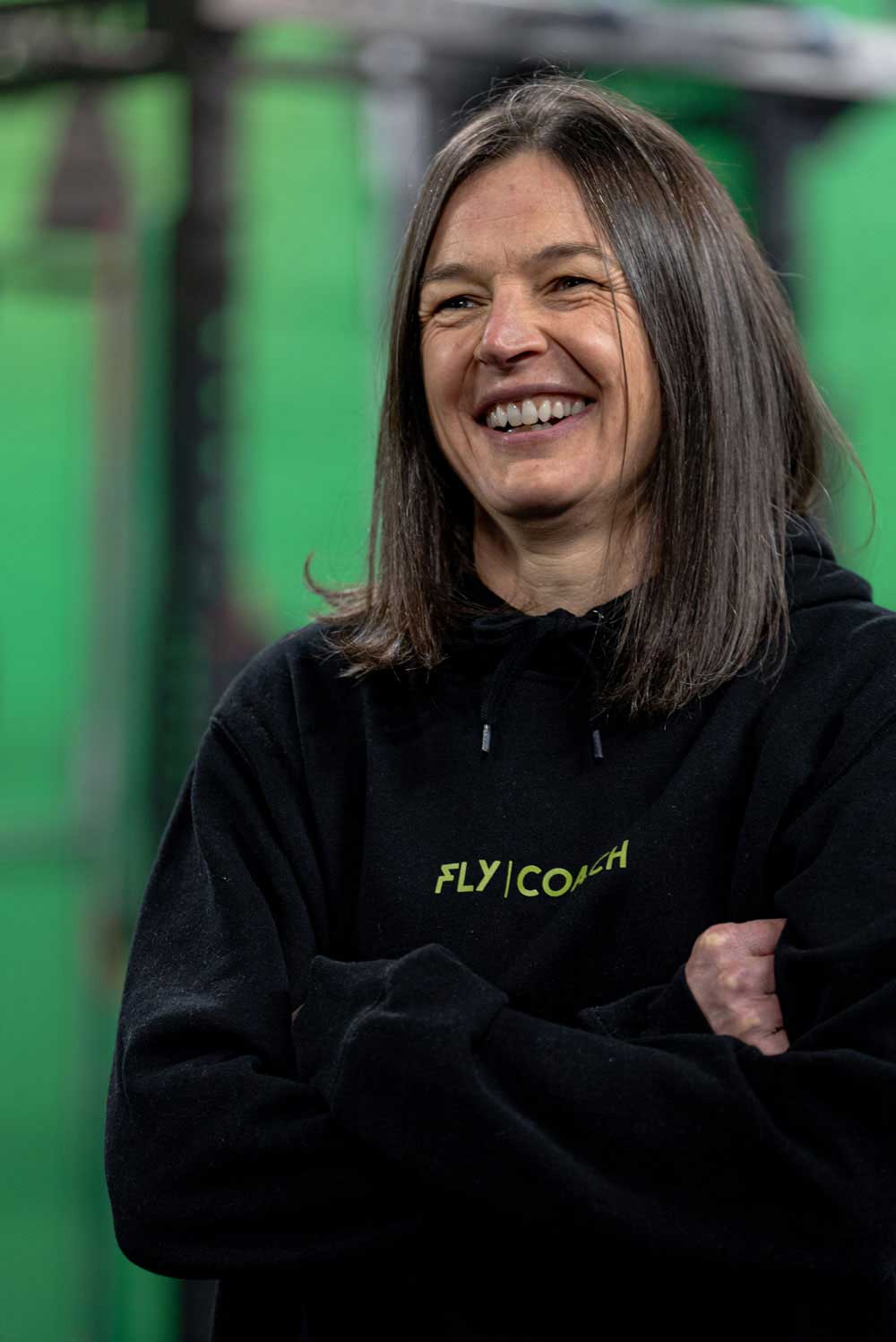 Smiling female coach in a black FLY COACH hoodie standing confidently in the gym